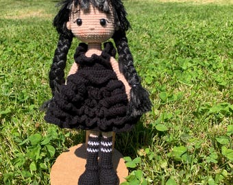 May include: A handmade crochet doll with black hair in two braids, wearing a black dress and black boots with white and black striped socks. The doll is standing on a round, light brown base, set against a green grassy background.