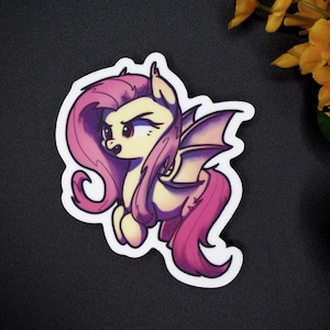 May include: A die-cut sticker featuring a cartoon pony with a pale yellow body, long flowing pink hair, and purple bat-like wings. The character has a determined expression with narrowed eyes and a small mouth, rendered in a vibrant, stylised art style.