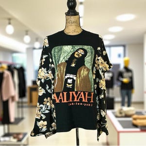 May include: Black graphic t-shirt featuring a portrait of Aaliyah with the text "AALIYAH" in orange. The sleeves are black with a floral pattern in cream and tan.