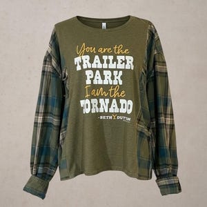 May include: An olive green long-sleeve shirt with plaid sleeves in shades of green, blue, and tan. The front features the text: You are the TRAILER PARK I am the TORNADO -BETH DUTTON in a mix of yellow script and bold white block lettering.