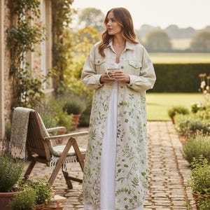May include: A long, beige denim jacket with a floral pattern on the lower half. The jacket is buttoned and worn over a white dress. The scene is set outdoors with a brick pathway and greenery.