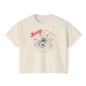May include: Off-white t-shirt featuring a spider web design with a spider and a figure, and the text "Sorry, I'm not home right now". The t-shirt has a round neck and short sleeves.
