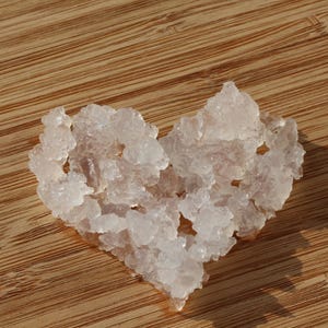 Può includere: Una collezione di grappoli traslucidi, irregolari, di colore bianco e rosa pallido, disposti a forma di cuore. I singoli pezzi presentano una consistenza ruvida e frastagliata e appaiono semitrasparenti, riflettendo la luce sulle superfici.
