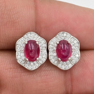 May include: A pair of stud earrings with oval, deep red gemstones, encircled by small, clear gemstones. The earrings have a floral design and are set in a silver-coloured metal.