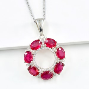 May include: A silver pendant necklace featuring a circular design adorned with oval-cut, vibrant red gemstones. The gemstones are set around a central open space, with smaller clear stones adding sparkle. The necklace hangs on a delicate silver chain.