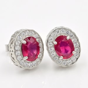 May include: A pair of silver stud earrings featuring oval-cut, vibrant red gemstones surrounded by a halo of small, clear gemstones. The earrings have a detailed, openwork design around the central stones.