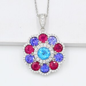 Puede incluir: Un collar con colgante en tono plateado que presenta una piedra preciosa redonda de color azul brillante en el centro, rodeada por un arreglo floral de piedras redondas rojas y moradas alternadas, enmarcado por pequeñas piedras blancas.
