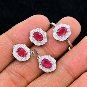 May include: A jewellery set consisting of a ring, a pendant, and two earrings, each featuring a central oval-cut red ruby surrounded by a halo of small white stones set in a silver-toned metal frame with a scalloped, flower-like edge design.