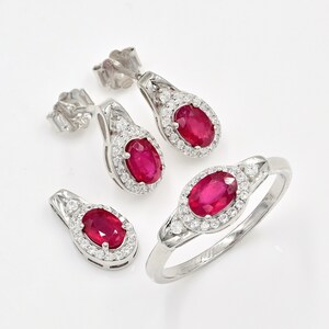 May include: A jewellery set consisting of a ring, a pendant, and a pair of stud earrings. Each piece features a central oval-cut red ruby surrounded by a halo of small white round-cut stones, all set in a polished silver-toned metal band and frame.