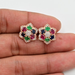 Könnte beinhalten: Ein Paar roségoldene Ohrringe in Blumenform mit einem Cluster aus bunten Edelsteinen. Die Ohrringe haben einen zentralen weißen Stein, der von grünen, blauen und roten Edelsteinen umgeben ist.