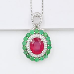 Könnte beinhalten: Eine silberne Halskette mit einem Anhänger, der einen großen ovalen Rubin-Edelstein zeigt, der von kleineren Smaragd- und weißen Edelsteinen umgeben ist.