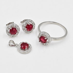 Peut inclure: Un ensemble de bijoux composé d'une bague, d'un pendentif et d'une paire de boucles d'oreilles à clous. Chaque pièce présente une pierre centrale ronde rouge foncé entourée d'un halo de petites pierres claires scintillantes sur métal argenté.