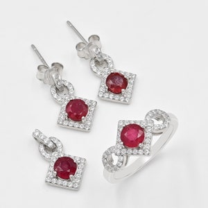 May include: A jewellery set consisting of two drop earrings, a pendant, and a ring, all crafted from silver-toned metal. Each piece features a central round red ruby stone set within a square frame encrusted with small white cubic zirconia stones.