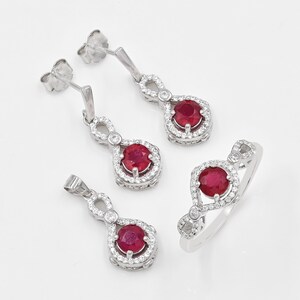 May include: A jewellery set consisting of a pair of drop earrings, a pendant, and a ring. Each piece features a round, deep red ruby centre stone surrounded by a halo of small white stones, set in a silver-toned metal with an infinity-style design.