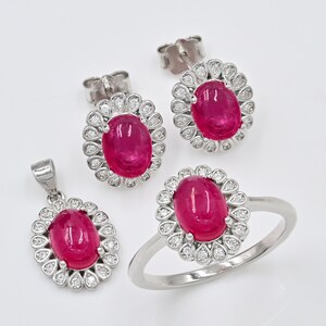 Op de afbeelding: Een sieradenset bestaande uit een ring, een hanger en een paar oorknopjes. Elk sieraad heeft een centrale ovale roze robijn in cabochon-slijpvorm, omringd door een halo van kleine witte diamanten gezet in gepolijst zilverkleurig metaal.