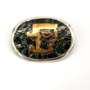 May include: A silver oval brooch with a green and gold mosaic design. The mosaic features a stylized letter 'F' in gold.