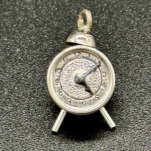 Może przedstawiać: Srebrny charm w kształcie budzika. Tarcza zegara ma czarne rzymskie cyfry godzinowe i czarne wskazówki. Charm ma pętelkę na górze do przymocowania do bransoletki lub naszyjnika.