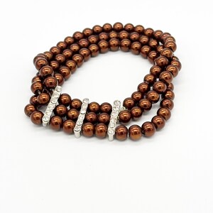 Könnte beinhalten: Ein dreireihiges Armband mit glänzenden, braunen, kugelförmigen Perlen. Das Armband ist mit drei silberfarbenen Stäben verziert, die jeweils mit kleinen, klaren, funkelnden Steinen besetzt sind. Der Schmuck ist vor weißem Hintergrund platziert.