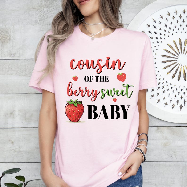 May include: A light pink t-shirt with the text "cousin of the berry sweet BABY" in red, green, and black. The design includes strawberry illustrations. The shirt is short-sleeved.