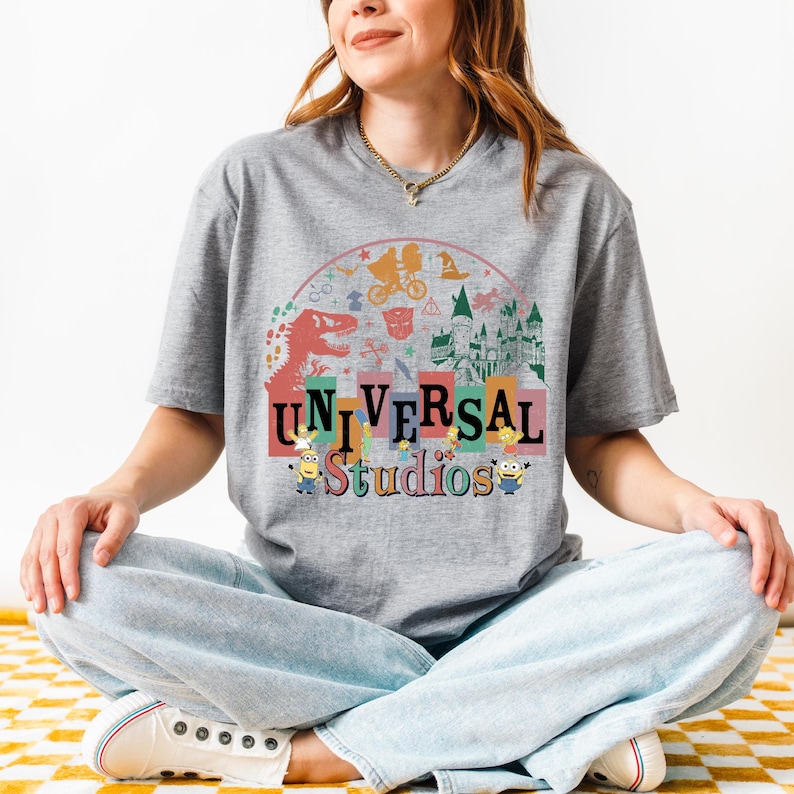 May include: A grey t-shirt with the words "UNIVERSAL Studios" in colourful block letters. The design includes illustrations of a dinosaur, Minions, and other theme park icons, set against a rainbow arch.