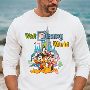 Op de afbeelding: Witte sweatshirt met een kleurrijke afbeelding van Disney-personages, waaronder Mickey Mouse, Minnie Mouse, Donald Duck en Goofy. De afbeelding bevat ook de tekst "Walt Disney World" en een kasteelillustratie.