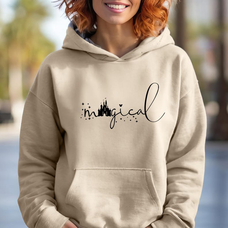 Puede incluir: Sudadera con capucha beige con bolsillo delantero. La palabra "magical" est&aacute; escrita en cursiva, con una silueta de castillo y estrellas. La sudadera es de color neutro.