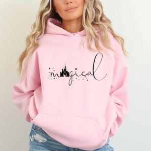 Puede incluir: Sudadera con capucha rosa claro con bolsillo delantero. La sudadera presenta un gr&aacute;fico negro de un castillo y la palabra "magical" en una fuente de escritura. El dise&ntilde;o est&aacute; rodeado de peque&ntilde;as estrellas.