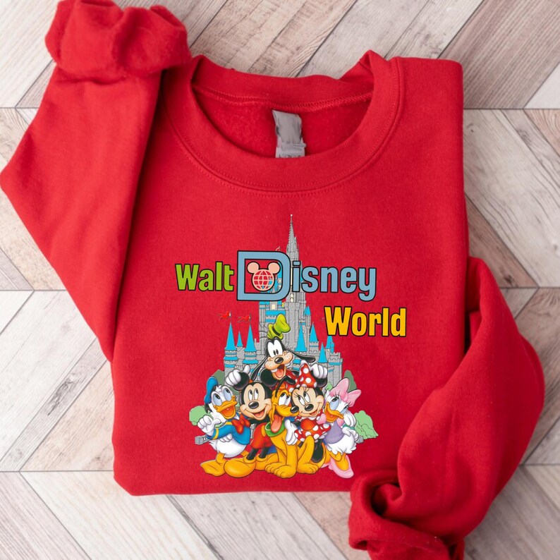 Op de afbeelding: Een rode sweatshirt met de tekst "Walt Disney World" in groen en geel, een kasteelafbeelding en stripfiguren zoals Mickey Mouse, Donald Duck en Goofy. De sweatshirt is gemaakt van een zacht materiaal.