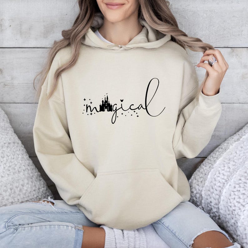 Puede incluir: Una sudadera con capucha beige con un bolsillo delantero y una capucha. La palabra "magical" est&aacute; escrita en cursiva, con una silueta de castillo y estrellas encima de la palabra.