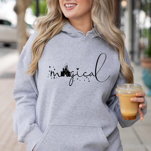 Puede incluir: Sudadera con capucha gris claro con un dise&ntilde;o negro que dice "magical" en cursiva. La palabra "magical" est&aacute; acompa&ntilde;ada de una silueta de castillo y estrellas. La sudadera tiene un bolsillo delantero y una capucha.