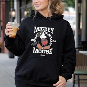 Puede incluir: Sudadera con capucha negra con un gr&aacute;fico blanco de Mickey Mouse con un atuendo rojo y blanco. El gr&aacute;fico incluye el texto "Mickey Mouse" y "1928" con un fondo a cuadros.