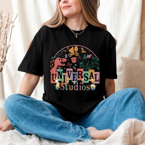 May include: Black t-shirt with a vibrant Universal Studios graphic, showcasing theme park icons like a dinosaur and Minions. The design incorporates imagery from popular films. The shirt is paired with blue jeans.