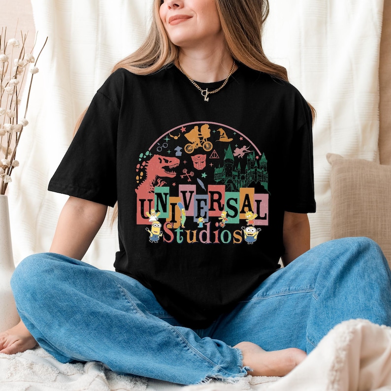 May include: Black t-shirt with a vibrant Universal Studios graphic, showcasing theme park icons like a dinosaur and Minions. The design incorporates imagery from popular films. The shirt is paired with blue jeans.