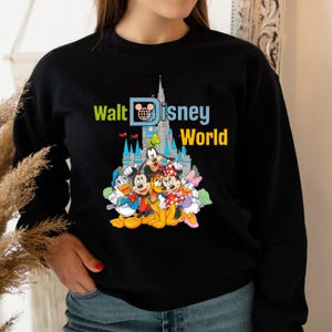 Op de afbeelding: Zwarte sweatshirt met een kleurrijke afbeelding van Disney-personages, waaronder Mickey Mouse, Donald Duck en Goofy, met de tekst "Walt Disney World" en een kasteelillustratie. Het ontwerp is levendig en opvallend.