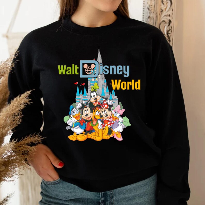 Op de afbeelding: Zwarte sweatshirt met een kleurrijke afbeelding van Disney-personages, waaronder Mickey Mouse, Donald Duck en Goofy, met de tekst "Walt Disney World" en een kasteelillustratie. Het ontwerp is levendig en opvallend.