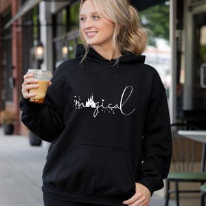 Puede incluir: Sudadera con capucha negra con un gr&aacute;fico blanco que dice "magical" en fuente cursiva, con una silueta de castillo y estrellas. La sudadera tiene un bolsillo delantero y una capucha con cord&oacute;n. La persona sostiene una taza de pl&aacute;stico transparente con una bebida marr&oacute;n.