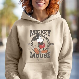 Puede incluir: Una sudadera con capucha beige con un gr&aacute;fico de Mickey Mouse de estilo vintage. El gr&aacute;fico presenta a Mickey Mouse con un atuendo rojo y negro, de pie en un c&iacute;rculo con el texto "Mickey Mouse" y los a&ntilde;os "1928" y "1928". El texto "Disney" tambi&eacute;n est&aacute; incluido en el gr&aacute;fico.