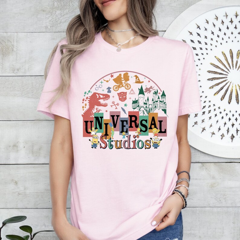 May include: A pale pink t-shirt with a vibrant Universal Studios graphic. The design incorporates a dinosaur, a castle, and Minions, with the words "UNIVERSAL Studios" in a fun font. The t-shirt has short sleeves.