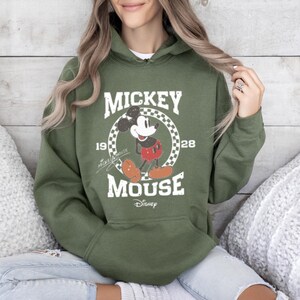 Puede incluir: Una sudadera con capucha verde con un gr&aacute;fico vintage de Mickey Mouse. El gr&aacute;fico presenta a Mickey Mouse en un c&iacute;rculo a cuadros con el texto "Mickey Mouse" y "1928" arriba y abajo del c&iacute;rculo. El texto "Disney" est&aacute; debajo del gr&aacute;fico.