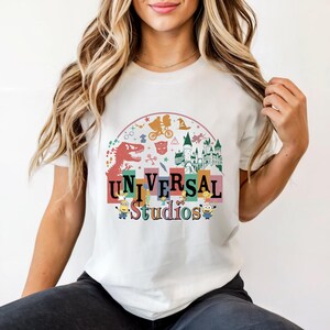 May include: White crew neck t-shirt with a vibrant Universal Studios graphic. The design incorporates a dinosaur, castle, and Minions, alongside the text 'Universal Studios'. The shirt is a casual wear item.