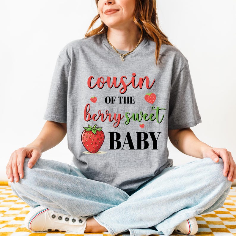 May include: A gray t-shirt with the text "Cousin of the berry sweet BABY" in red, green, and black. The design includes a strawberry graphic and small heart accents. The shirt is worn by a person.