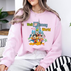Op de afbeelding: Een lichtroze sweatshirt met een kleurrijke afbeelding van Disney-figuren voor een kasteel. De tekst "Walt Disney World" staat in groen en geel boven de figuren.