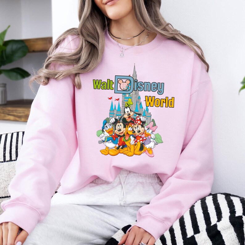 Op de afbeelding: Een lichtroze sweatshirt met een kleurrijke afbeelding van Disney-figuren voor een kasteel. De tekst "Walt Disney World" staat in groen en geel boven de figuren.