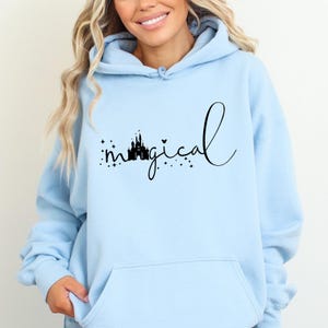 Puede incluir: Sudadera con capucha azul claro con la palabra "magical" en escritura negra, con una silueta de castillo. La sudadera con capucha tiene una capucha con cord&oacute;n y un bolsillo delantero. La modelo lleva la sudadera con capucha.