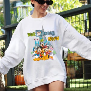 Op de afbeelding: Witte sweatshirt met een kleurrijke afbeelding van Disney-personages, waaronder Mickey Mouse, Donald Duck en Goofy, voor een kasteel. De tekst "Walt Disney World" staat in groen en geel boven de personages.