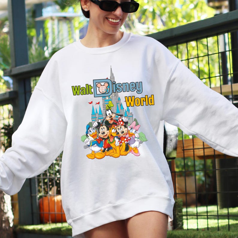Op de afbeelding: Witte sweatshirt met een kleurrijke afbeelding van Disney-personages, waaronder Mickey Mouse, Donald Duck en Goofy, voor een kasteel. De tekst "Walt Disney World" staat in groen en geel boven de personages.