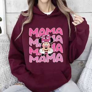 以下が含まれることがあります： 濃い赤紫色のフード付きスウェットシャツに、ピンク色のミニーマウスのグラフィックがプリントされています。ミニーマウスは眼鏡とピンクのリボンをつけています。グラフィックは4回繰り返され、ピンク色の文字で「MAMA」と書いてあります。