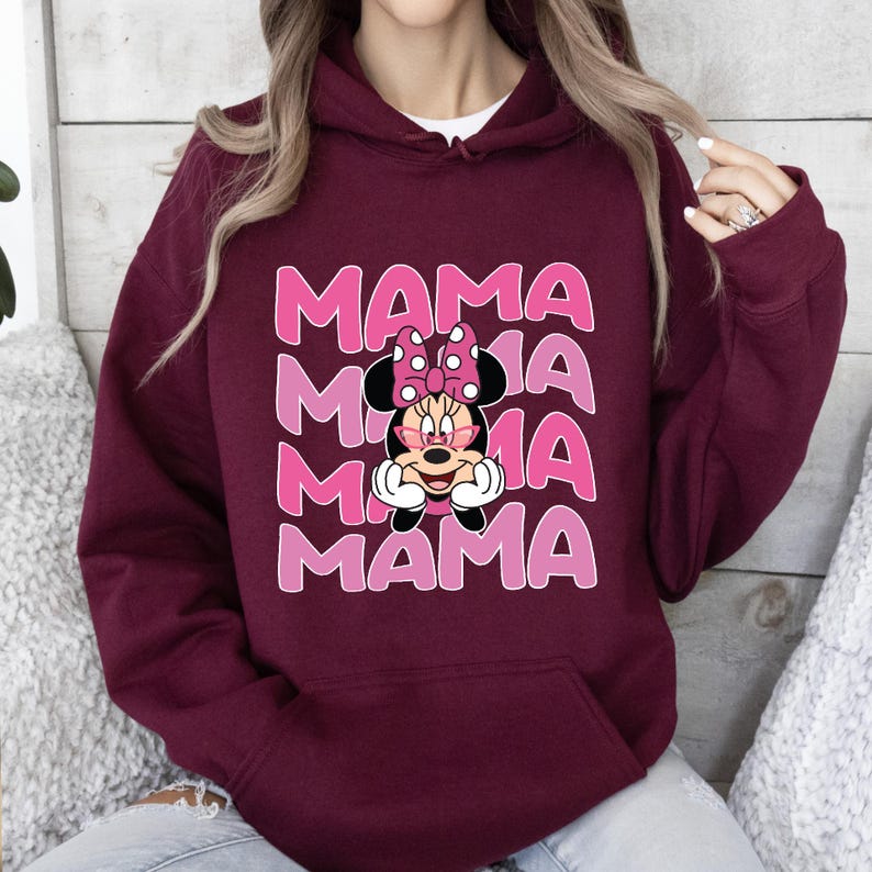 以下が含まれることがあります： 濃い赤紫色のフード付きスウェットシャツに、ピンク色のミニーマウスのグラフィックがプリントされています。ミニーマウスは眼鏡とピンクのリボンをつけています。グラフィックは4回繰り返され、ピンク色の文字で「MAMA」と書いてあります。