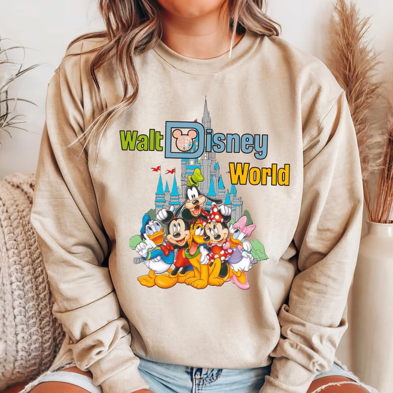 Op de afbeelding: Beige sweatshirt met een kleurrijke afbeelding van Disney-personages, waaronder Mickey Mouse en Donald Duck. De tekst "Walt Disney World" staat boven de personages. De sweatshirt heeft lange mouwen en is gemaakt van zacht materiaal.
