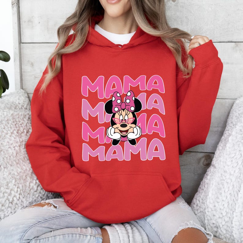 以下が含まれることがあります： 赤いフード付きスウェットシャツに、ピンクと白のミニーマウスのグラフィックがプリントされています。"MAMA"という文字が4回繰り返されています。